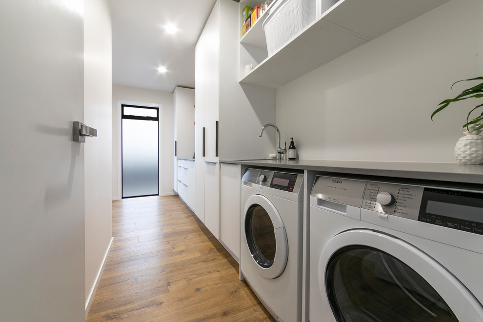 Large contemporary galley laundry room in Auckland with a single-bowl sink, flat-panel cabinets, white cabinets, quartz benchtops, white splashback, laminate floors, brown floor and grey benchtop.