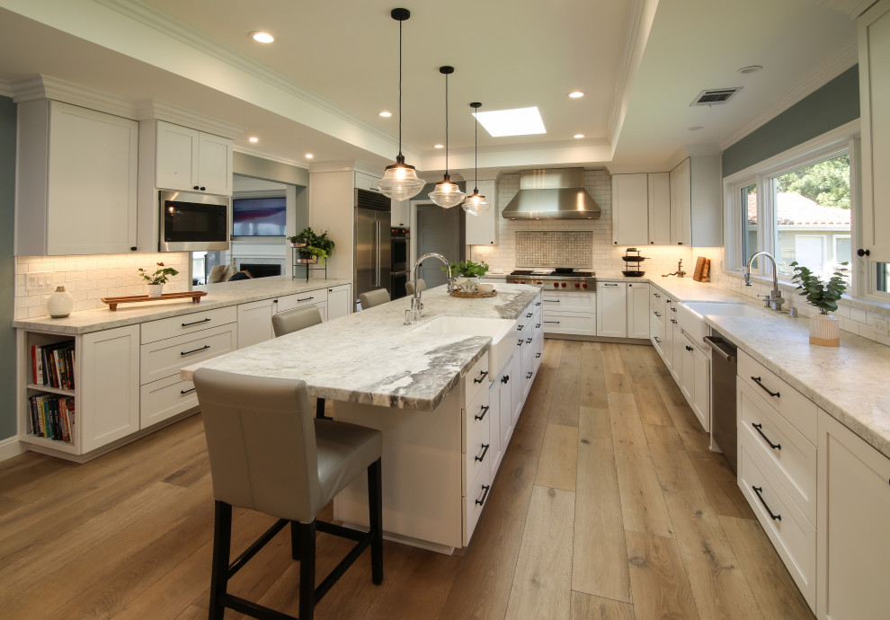 This is an example of a large country l-shaped eat-in kitchen in San Francisco with a farmhouse sink, quartzite benchtops, white splashback, stainless steel appliances, medium hardwood floors, white benchtop, shaker cabinets, white cabinets, ceramic splashback, with island, brown floor and recessed.