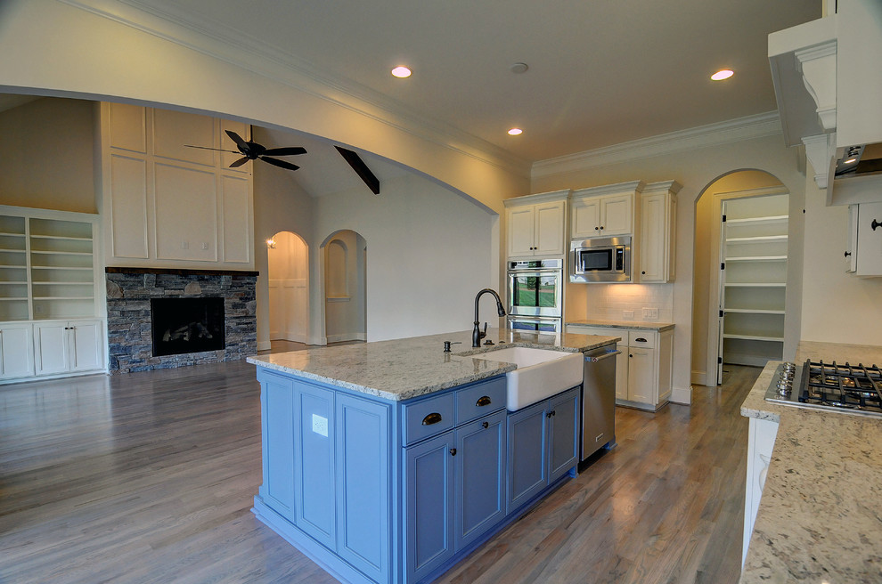 Large transitional l-shaped open plan kitchen in Charlotte with a farmhouse sink, recessed-panel cabinets, white cabinets, granite benchtops, white splashback, subway tile splashback, stainless steel appliances, light hardwood floors and with island.