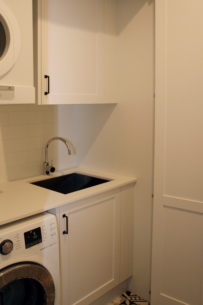 Photo of a mid-sized traditional single-wall utility room in Sydney with an undermount sink, shaker cabinets, white cabinets, solid surface benchtops, white walls, ceramic floors, an integrated washer and dryer, multi-coloured floor and white benchtop.