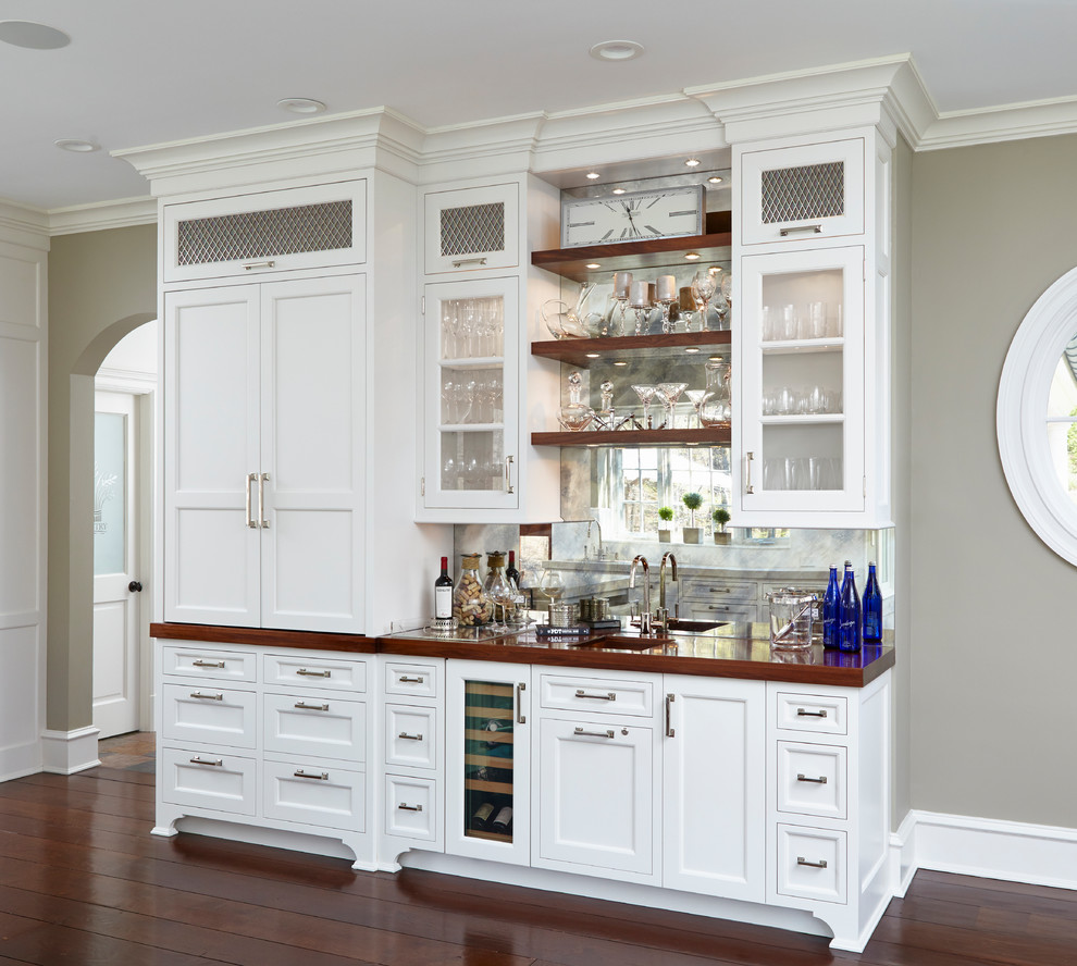 Mid-sized traditional single-wall wet bar in New York with an undermount sink, glass-front cabinets, white cabinets, wood benchtops, mirror splashback, dark hardwood floors, brown floor and brown benchtop.