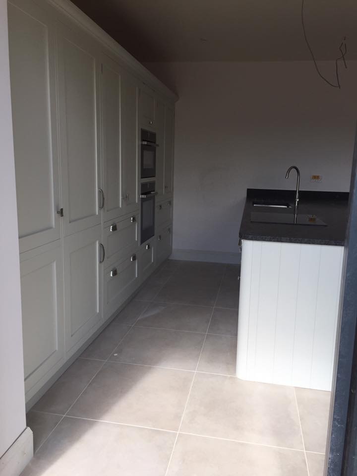 Photo of a large contemporary single-wall open plan kitchen in Wiltshire with a drop-in sink, flat-panel cabinets, white cabinets, granite benchtops, white splashback, cement tile splashback, black appliances, ceramic floors, with island and beige floor.