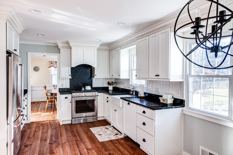 Photo of a mid-sized country l-shaped eat-in kitchen in New York with a farmhouse sink, shaker cabinets, white cabinets, white splashback, stainless steel appliances, no island and black benchtop.