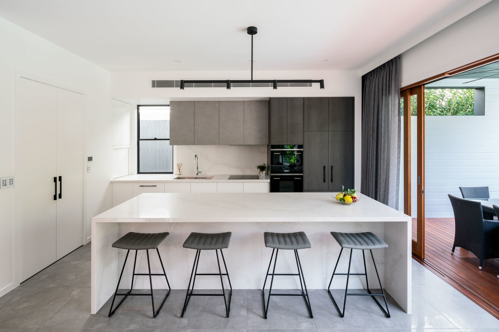 This is an example of a contemporary galley kitchen in Brisbane with a single-bowl sink, flat-panel cabinets, white cabinets, white splashback, stone slab splashback, panelled appliances, with island, grey floor and white benchtop.