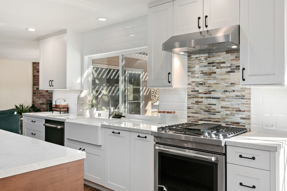 This is an example of a large transitional l-shaped open plan kitchen in San Diego with a farmhouse sink, shaker cabinets, white cabinets, quartz benchtops, multi-coloured splashback, ceramic splashback, stainless steel appliances, vinyl floors, with island, brown floor and grey benchtop.