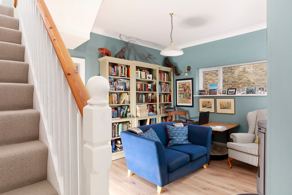 This is an example of a mid-sized eclectic home office in Kent with a library, blue walls, light hardwood floors, a wood stove, a stone fireplace surround and beige floor.
