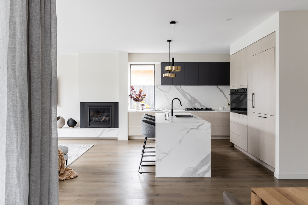 This is an example of a contemporary open plan kitchen in Melbourne with a double-bowl sink, flat-panel cabinets, beige cabinets, white splashback, black appliances, medium hardwood floors, with island and white benchtop.