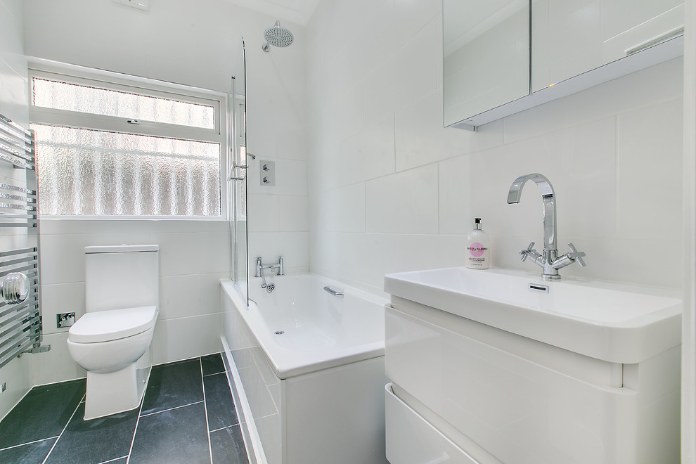 This is an example of a mid-sized transitional kids bathroom in London with a drop-in sink, furniture-like cabinets, white cabinets, solid surface benchtops, a drop-in tub, a shower/bathtub combo, a two-piece toilet, white tile, ceramic tile, white walls and ceramic floors.