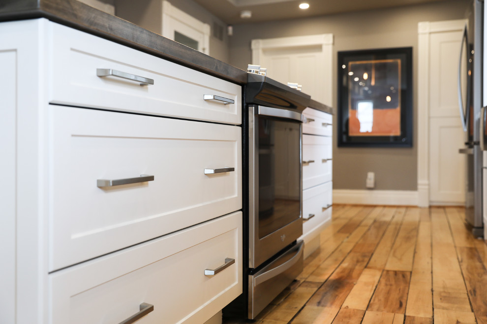 Mid-sized transitional single-wall medium tone wood floor and brown floor eat-in kitchen photo with a farmhouse sink, shaker cabinets, white cabinets, white backsplash, stainless steel appliances, an island and white countertops