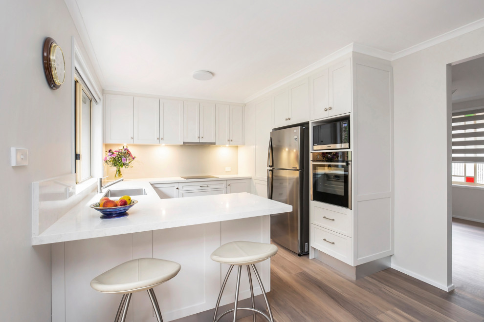 This is an example of a medium sized modern u-shaped kitchen/diner in Sydney with a submerged sink, shaker cabinets, white cabinets, engineered stone countertops, white splashback, engineered quartz splashback, stainless steel appliances, light hardwood flooring, no island and white worktops.