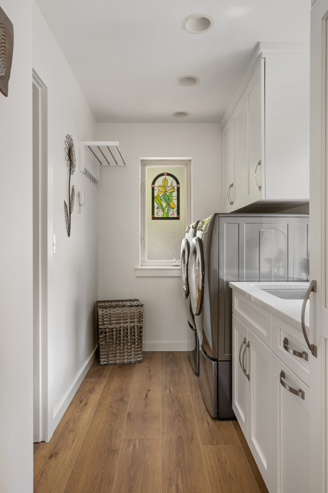 Photo of a mid-sized beach style single-wall dedicated laundry room in Seattle with a drop-in sink, beaded inset cabinets, white cabinets, marble benchtops, white splashback, white walls, vinyl floors, a side-by-side washer and dryer, brown floor, white benchtop and coffered.
