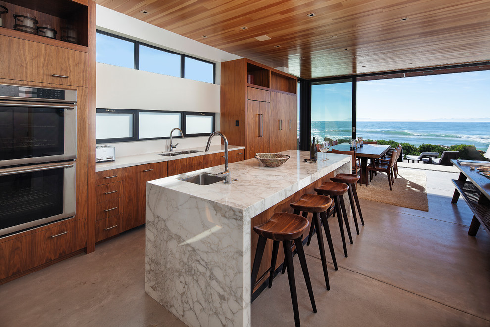 Photo of a mid-sized beach style open plan kitchen in Los Angeles with an undermount sink, flat-panel cabinets, medium wood cabinets, marble benchtops, panelled appliances, concrete floors, with island and grey floor.