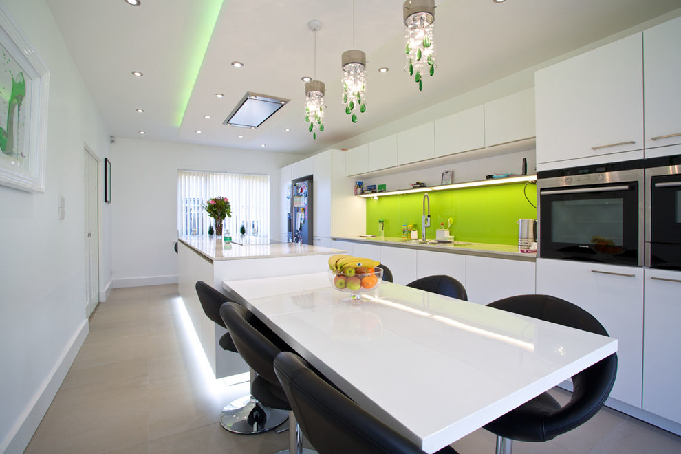 Large contemporary galley open plan kitchen in Hampshire with an integrated sink, flat-panel cabinets, white cabinets, solid surface benchtops, green splashback, glass sheet splashback, stainless steel appliances, porcelain floors and with island.