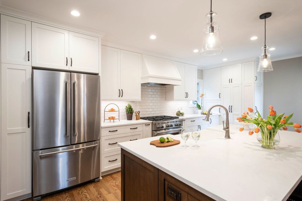 This is an example of a transitional u-shaped eat-in kitchen in Indianapolis with a farmhouse sink, shaker cabinets, white cabinets, quartz benchtops, white splashback, ceramic splashback, stainless steel appliances, medium hardwood floors, with island, brown floor and white benchtop.