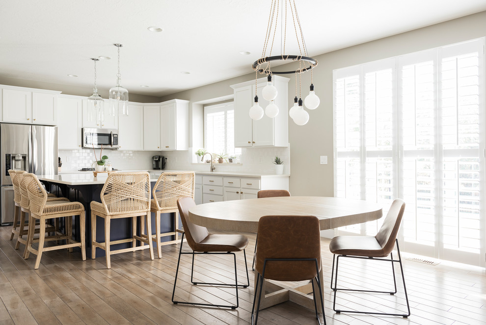 This is an example of a beach style l-shaped eat-in kitchen in Denver with shaker cabinets, white cabinets, white splashback, subway tile splashback, stainless steel appliances, with island, white benchtop and medium hardwood floors.