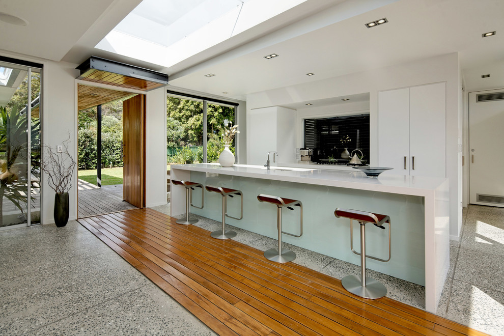 Inspiration for a mid-sized beach style u-shaped separate kitchen in Auckland with a drop-in sink, white cabinets, granite benchtops, black splashback, ceramic splashback, stainless steel appliances, concrete floors, with island and flat-panel cabinets.