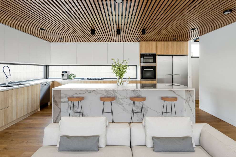Photo of a large contemporary l-shaped open plan kitchen in Melbourne with an undermount sink, flat-panel cabinets, light wood cabinets, marble benchtops, window splashback, black appliances, light hardwood floors, with island, brown floor, white benchtop and timber.