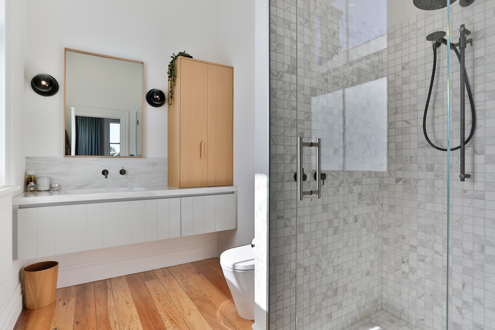 This is an example of a mid-sized scandinavian master bathroom in Auckland with grey cabinets, an alcove shower, a one-piece toilet, gray tile, marble, white walls, medium hardwood floors, marble benchtops, brown floor, a hinged shower door, white benchtops and an integrated sink.
