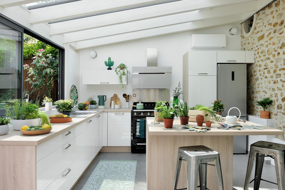 Design ideas for a contemporary l-shaped eat-in kitchen with a drop-in sink, flat-panel cabinets, white cabinets, wood benchtops, white splashback, metal splashback, black appliances, concrete floors, with island, grey floor and beige benchtop.