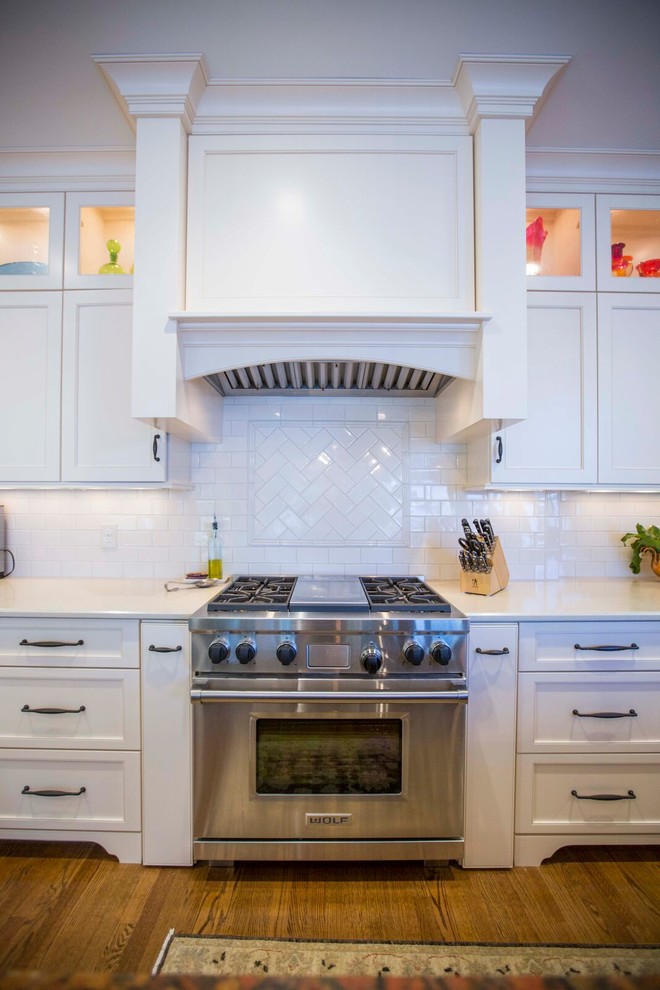 Inspiration for a large transitional l-shaped light wood floor and brown floor enclosed kitchen remodel with a farmhouse sink, shaker cabinets, white cabinets, solid surface countertops, white backsplash, porcelain backsplash, stainless steel appliances and an island