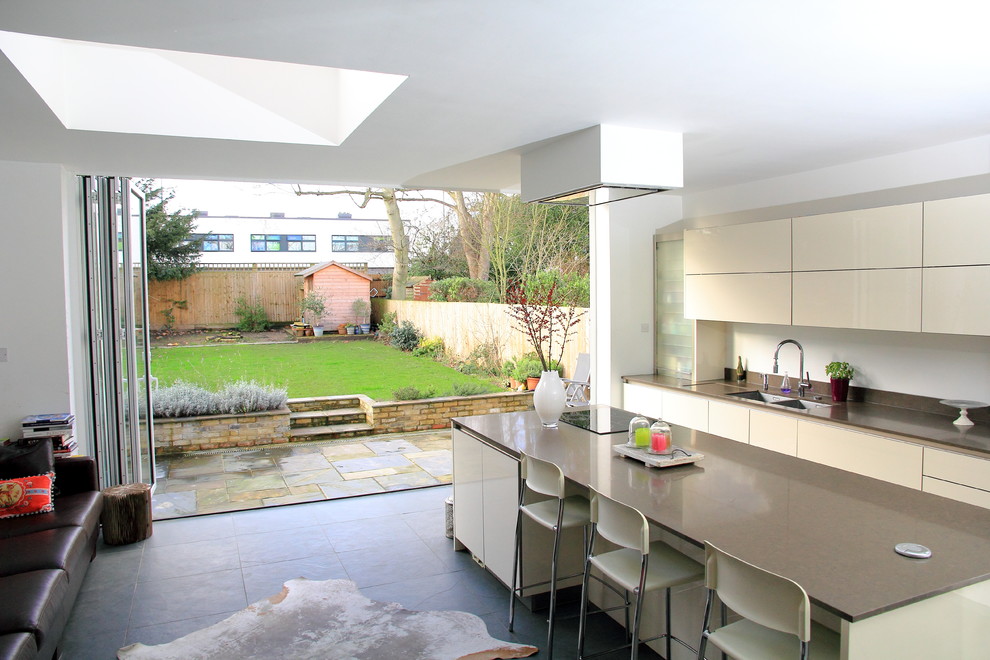 This is an example of a large modern kitchen/diner in London with flat-panel cabinets, white cabinets, white splashback, ceramic splashback, integrated appliances, travertine flooring and an island.