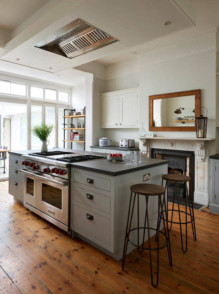 Inspiration for a large transitional open plan kitchen in Essex with a farmhouse sink, shaker cabinets, grey cabinets, black splashback, stainless steel appliances, medium hardwood floors, with island, brown floor and black benchtop.