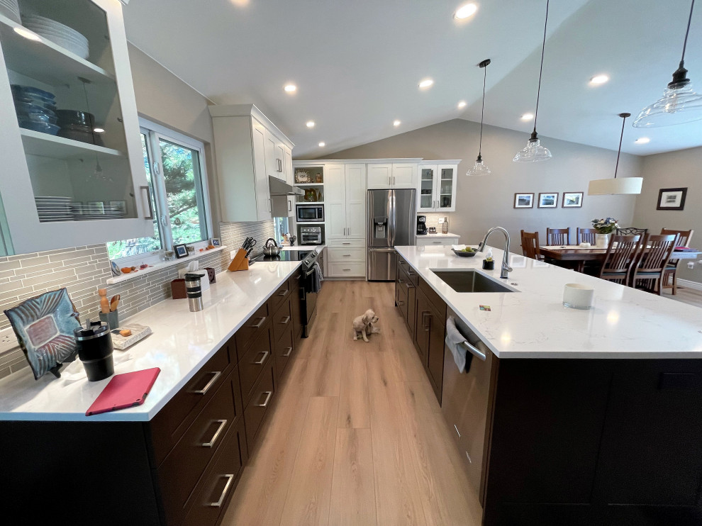 This is an example of a large traditional l-shaped open plan kitchen in Denver with a single-bowl sink, shaker cabinets, engineered stone countertops, grey splashback, glass sheet splashback, stainless steel appliances, vinyl flooring, an island, beige floors and a vaulted ceiling.