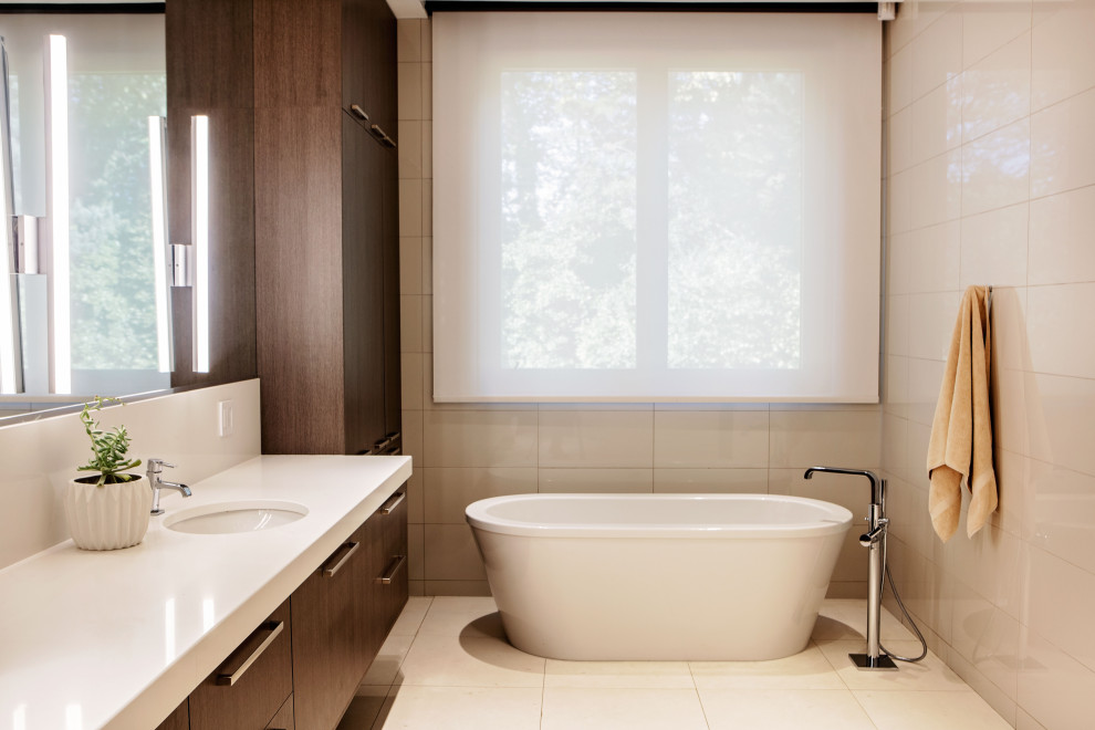 На фото: главная ванная комната среднего размера в стиле модернизм с плоскими фасадами, темными деревянными фасадами, отдельно стоящей ванной, душем в нише, унитазом-моноблоком, белой плиткой, керамогранитной плиткой, белыми стенами, полом из керамогранита, врезной раковиной, столешницей из искусственного кварца, белым полом, душем с распашными дверями, белой столешницей, сиденьем для душа, тумбой под две раковины и встроенной тумбой