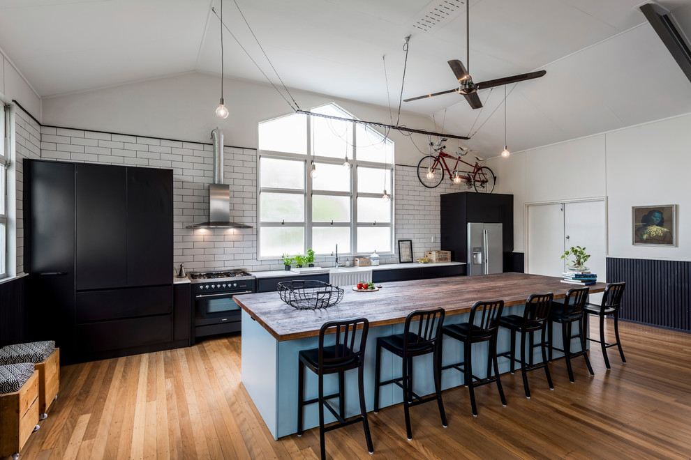 Large industrial galley open plan kitchen in Brisbane with a farmhouse sink, flat-panel cabinets, black cabinets, quartz benchtops, white splashback, stainless steel appliances, medium hardwood floors, with island, subway tile splashback and brown floor.