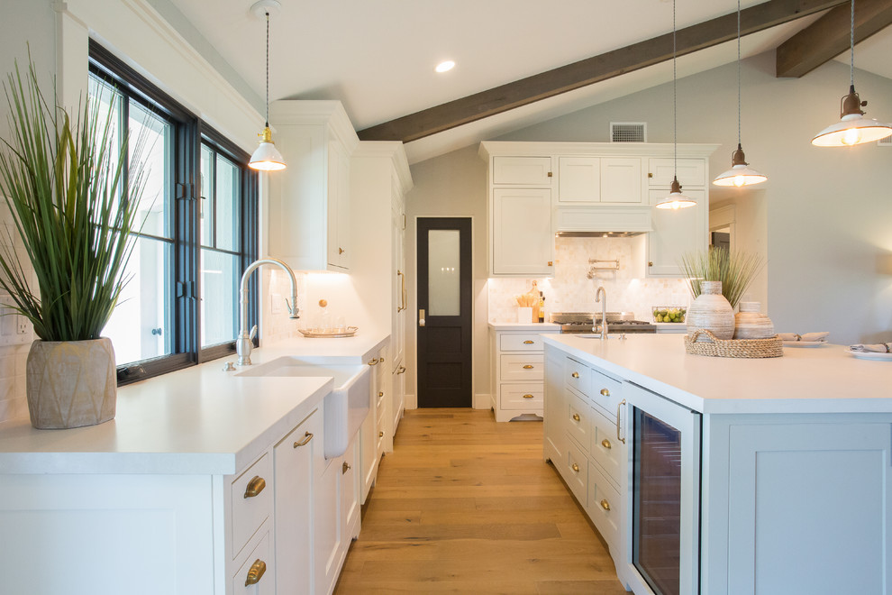 This is an example of a country l-shaped kitchen in Phoenix with a farmhouse sink, shaker cabinets, white cabinets, white splashback, mosaic tile splashback, panelled appliances, medium hardwood floors, with island, brown floor and white benchtop.