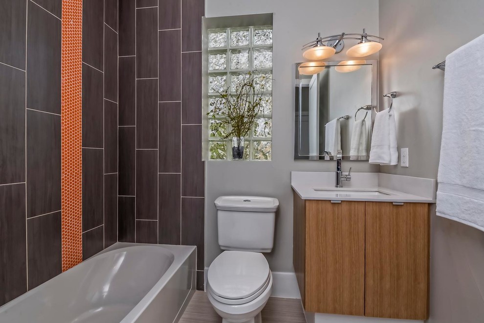 This is an example of a mid-sized contemporary 3/4 bathroom in Denver with flat-panel cabinets, medium wood cabinets, an alcove tub, a shower/bathtub combo, a two-piece toilet, brown tile, orange tile, porcelain tile, grey walls, plywood floors, an undermount sink and solid surface benchtops.
