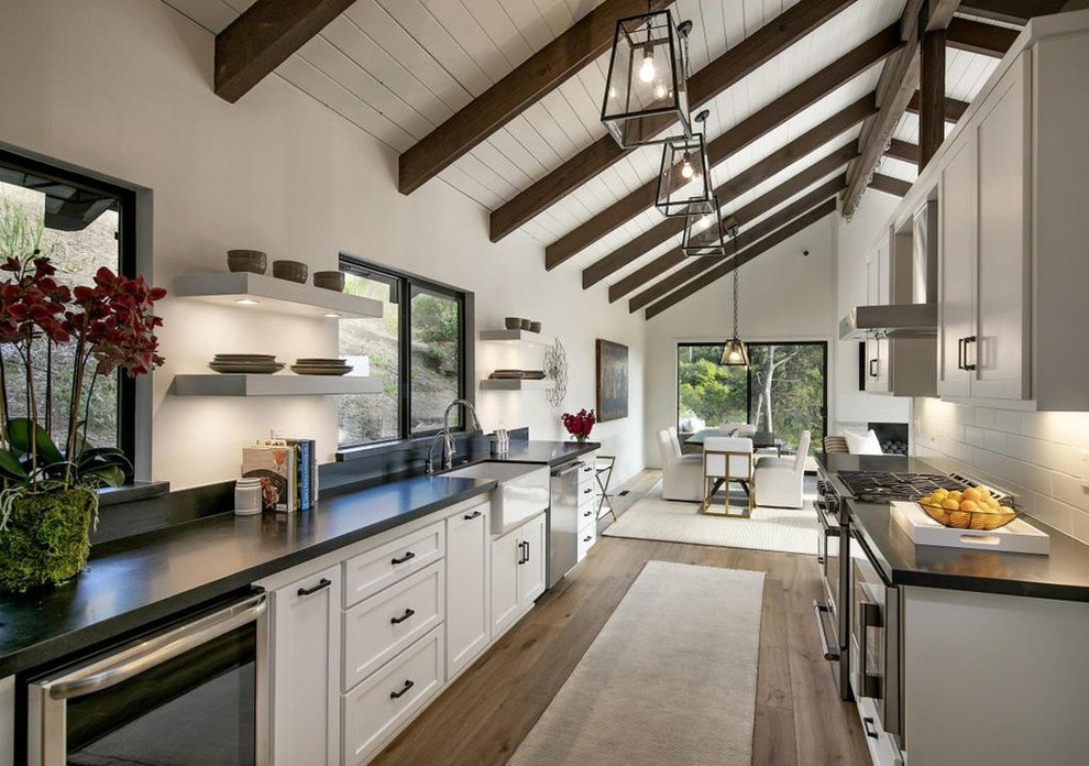 Inspiration for a mid-sized modern galley eat-in kitchen in Santa Barbara with a farmhouse sink, shaker cabinets, white cabinets, granite benchtops, white splashback, ceramic splashback, stainless steel appliances, medium hardwood floors, no island, brown floor and black benchtop.