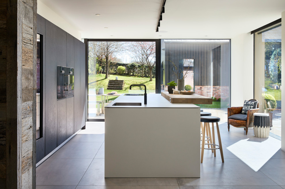 Contemporary grey and white single-wall open plan kitchen in Other with a built-in sink, flat-panel cabinets, white cabinets, laminate countertops, white splashback, black appliances, concrete flooring, an island, grey floors and white worktops.