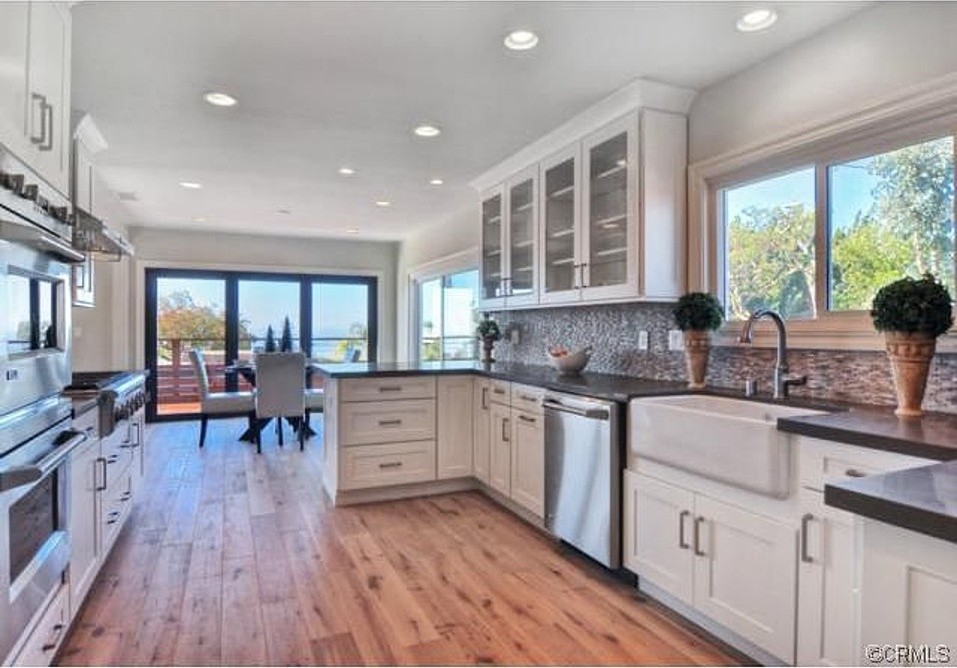Inspiration for a mid-sized beach style galley eat-in kitchen in Orange County with a farmhouse sink, shaker cabinets, white cabinets, quartz benchtops, multi-coloured splashback, glass tile splashback, stainless steel appliances, light hardwood floors and no island.