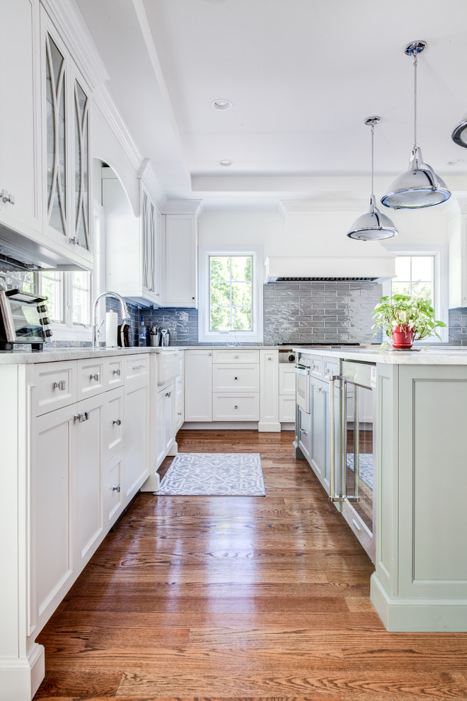 Design ideas for a mid-sized transitional u-shaped open plan kitchen in New York with a farmhouse sink, shaker cabinets, white cabinets, quartz benchtops, grey splashback, glass tile splashback, panelled appliances, light hardwood floors, with island and brown floor.