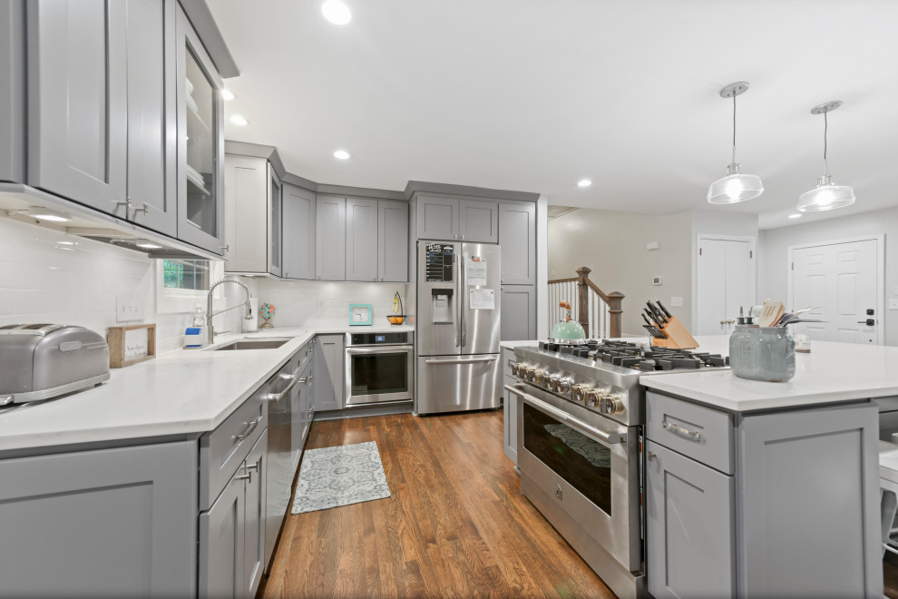 Photo of a large traditional galley eat-in kitchen in Atlanta with a farmhouse sink, raised-panel cabinets, grey cabinets, granite benchtops, white splashback, subway tile splashback, stainless steel appliances, medium hardwood floors, with island, brown floor and white benchtop.