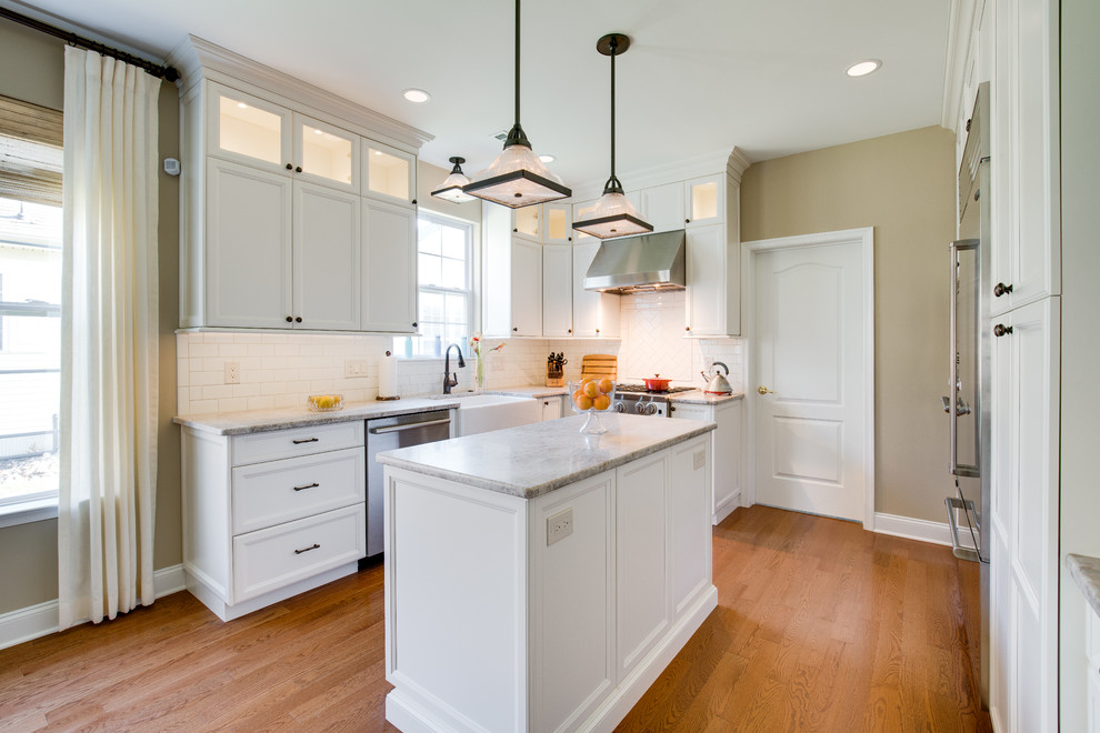 This is an example of a mid-sized traditional kitchen in Philadelphia with a farmhouse sink, recessed-panel cabinets, white cabinets, quartzite benchtops, white splashback, porcelain splashback, stainless steel appliances, medium hardwood floors, with island and brown floor.