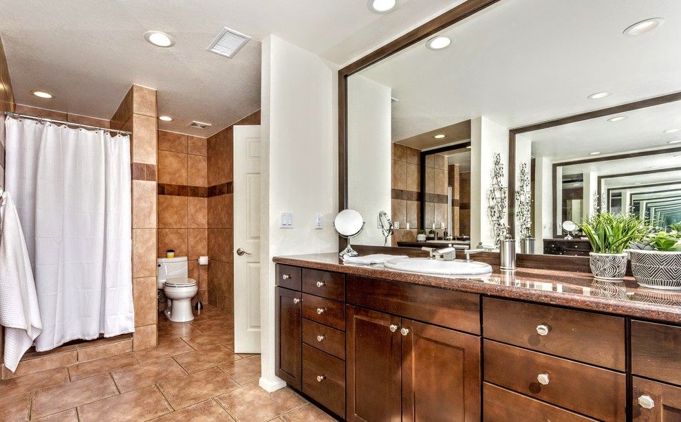 Photo of a large mediterranean master bathroom in Other with recessed-panel cabinets, dark wood cabinets, an alcove shower, a two-piece toilet, brown tile, ceramic tile, white walls, ceramic floors, a drop-in sink, granite benchtops, brown floor, a shower curtain and red benchtops.