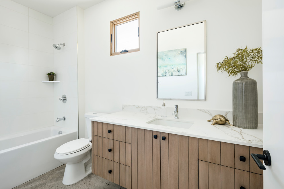 Photo of a bathroom in Phoenix with flat-panel cabinets, brown cabinets, an alcove tub, an alcove shower, a two-piece toilet, white tile, porcelain tile, white walls, concrete floors, a drop-in sink, engineered quartz benchtops, grey floor and a shower curtain.