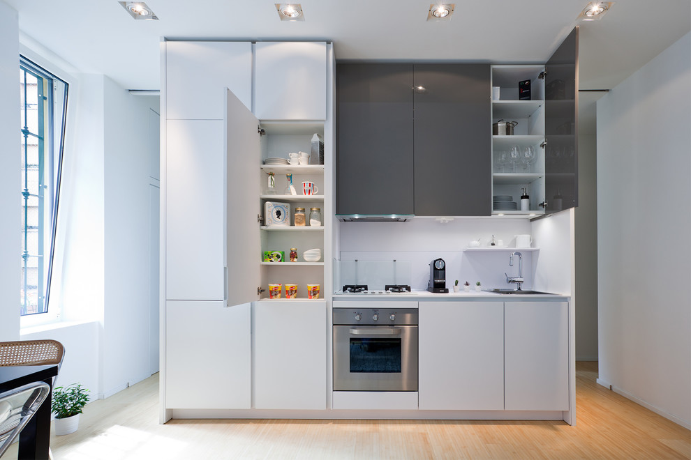 Photo of a small contemporary single-wall eat-in kitchen in Milan with a drop-in sink, flat-panel cabinets, white cabinets, glass tile splashback, stainless steel appliances and light hardwood floors.