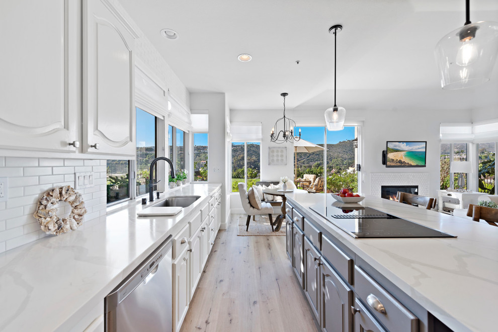 Design ideas for a mid-sized beach style single-wall eat-in kitchen in Orange County with an undermount sink, raised-panel cabinets, white cabinets, quartz benchtops, white splashback, ceramic splashback, stainless steel appliances, vinyl floors, with island, beige floor and white benchtop.
