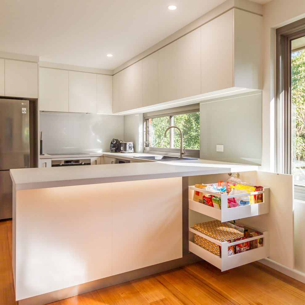 Mid-sized contemporary u-shaped eat-in kitchen in Melbourne with a drop-in sink, flat-panel cabinets, white cabinets, quartz benchtops, white splashback, glass sheet splashback, stainless steel appliances, medium hardwood floors, a peninsula, brown floor and white benchtop.