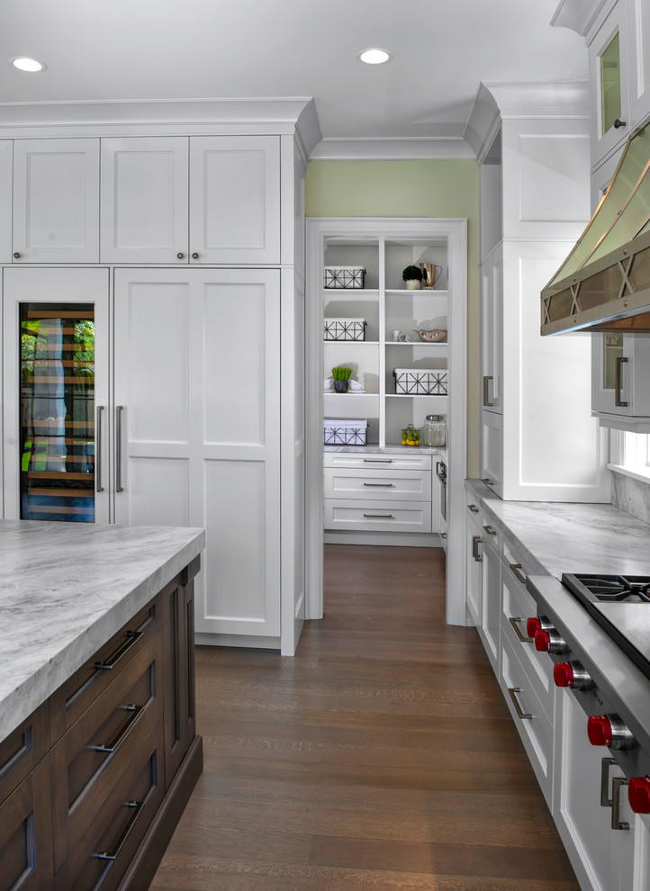This is an example of a large transitional kitchen pantry in Detroit with shaker cabinets, white cabinets, marble benchtops, marble splashback, stainless steel appliances, medium hardwood floors, with island and brown floor.
