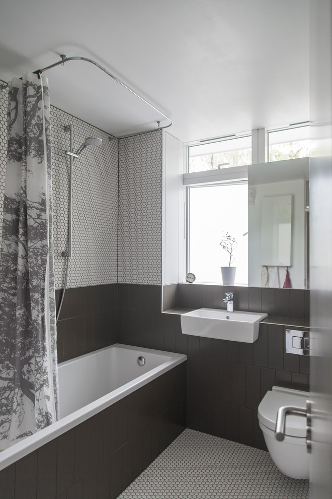 Small midcentury bathroom in London with glass-front cabinets, a drop-in tub, a shower/bathtub combo, a wall-mount toilet, gray tile, ceramic tile, white walls, ceramic floors, an integrated sink and tile benchtops.