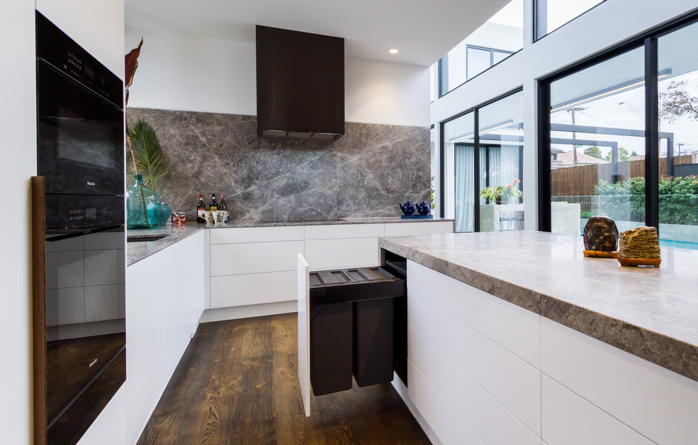 Photo of a large modern l-shaped open plan kitchen in Melbourne with an undermount sink, flat-panel cabinets, white cabinets, grey splashback, marble splashback, black appliances, medium hardwood floors, with island, brown floor, grey benchtop and limestone benchtops.