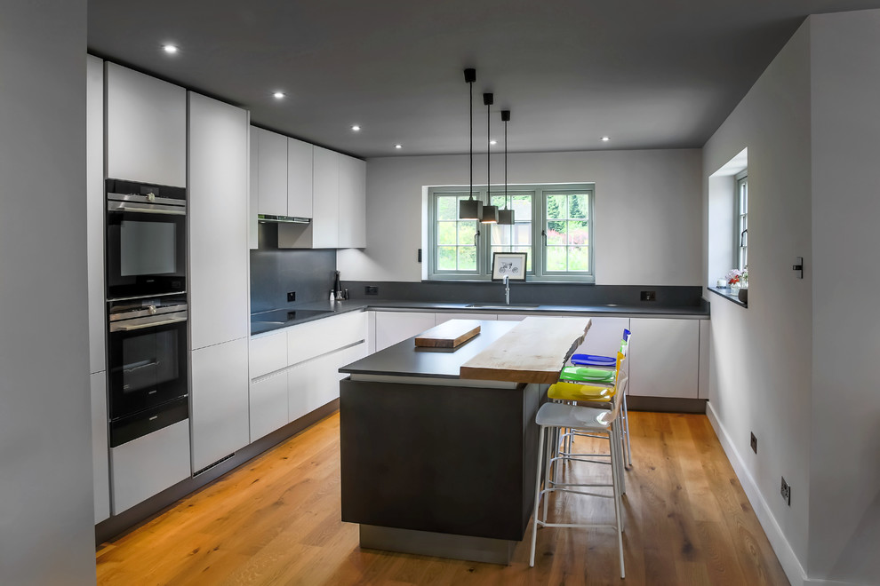 Mid-sized contemporary l-shaped kitchen in Essex with flat-panel cabinets, white cabinets, with island, black benchtop, black splashback, glass sheet splashback and medium hardwood floors.