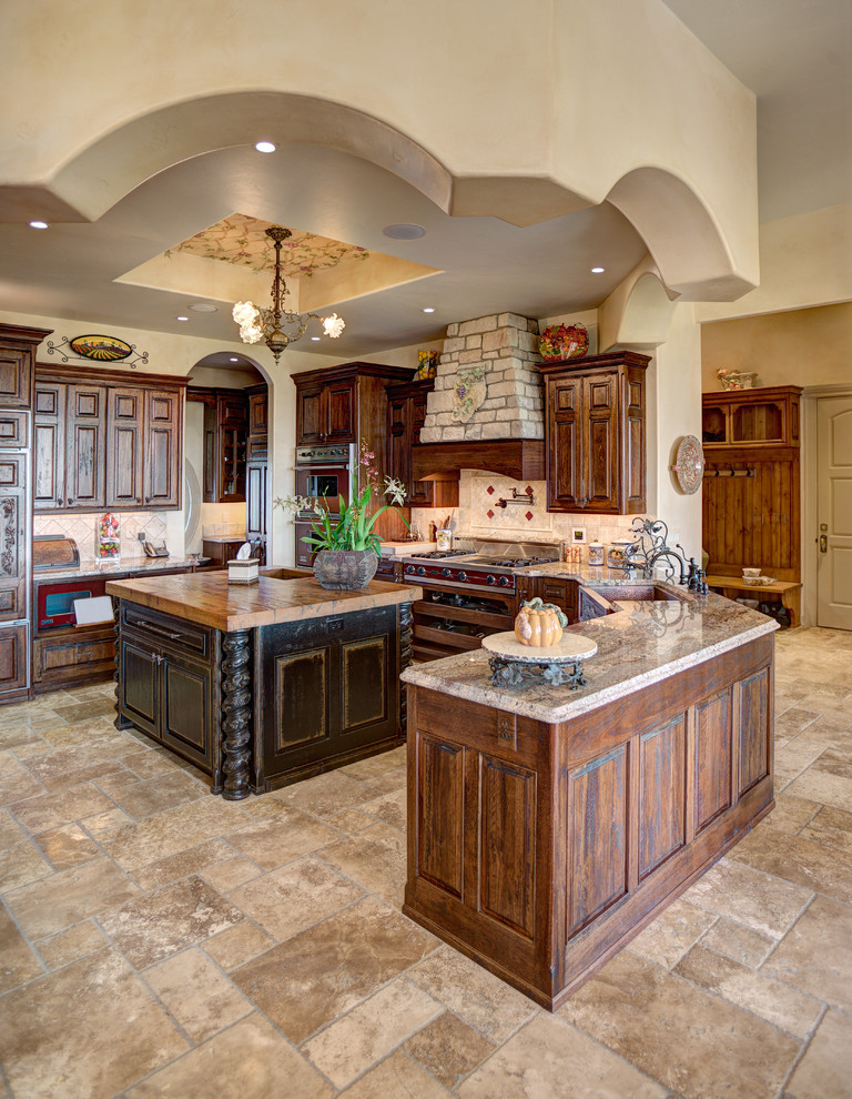 Example of a large tuscan u-shaped travertine floor and brown floor eat-in kitchen design with a farmhouse sink, raised-panel cabinets, dark wood cabinets, granite countertops, beige backsplash, stone tile backsplash and an island