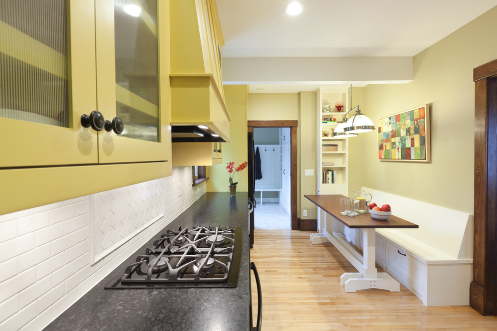 Photo of a mid-sized traditional l-shaped eat-in kitchen in Minneapolis with a farmhouse sink, yellow cabinets, granite benchtops, white splashback, ceramic splashback, black appliances, light hardwood floors, shaker cabinets and no island.