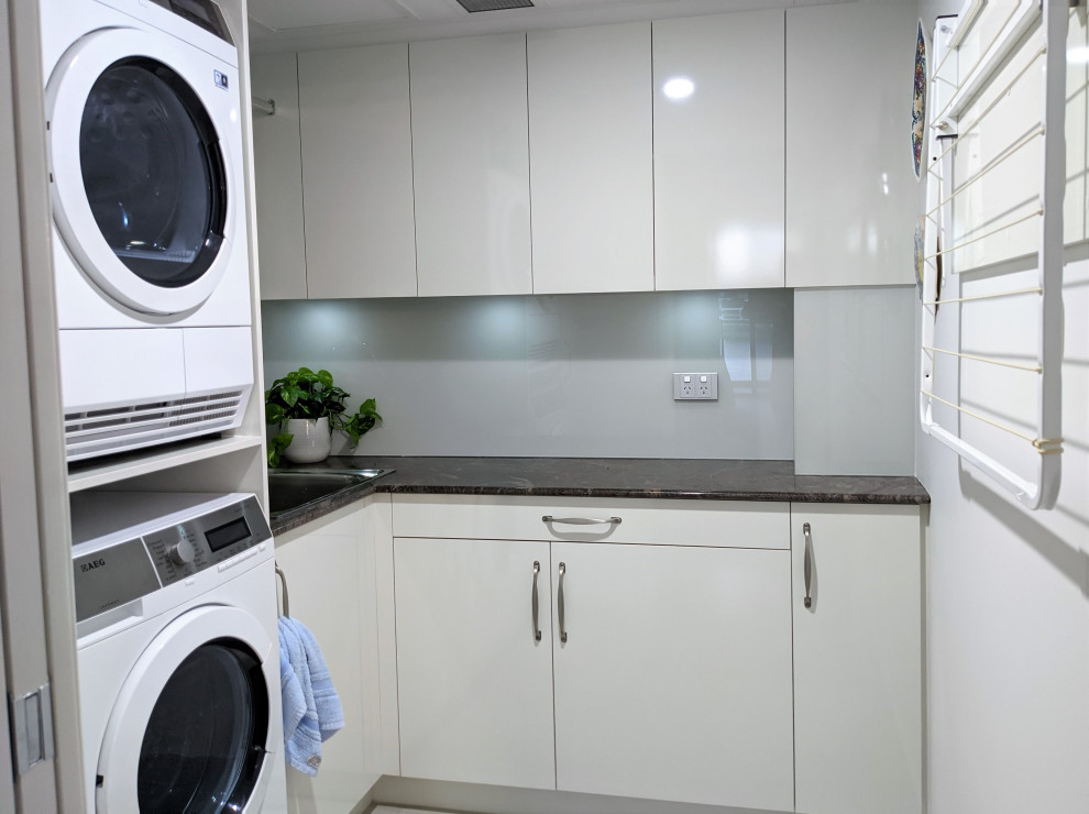 Photo of a small contemporary l-shaped dedicated laundry room in Brisbane with a drop-in sink, flat-panel cabinets, grey cabinets, granite benchtops, grey splashback, glass sheet splashback, white walls, a stacked washer and dryer and black benchtop.