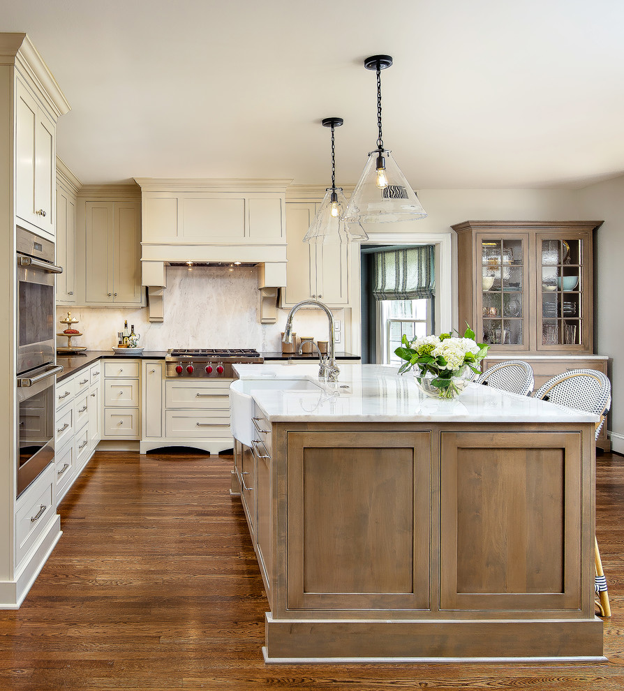 This is an example of a traditional l-shaped kitchen in New Orleans with a farmhouse sink, shaker cabinets, beige cabinets, stainless steel appliances, medium hardwood floors, with island, brown floor and black benchtop.